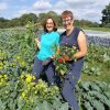 2 smiling volunteers standing in the sunshine in a field, in the process of weeding a bed of kale, holding large weeds