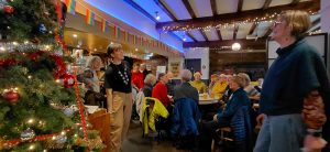 Group of singers mostly sat at pub tables singing beside a Christmas tree