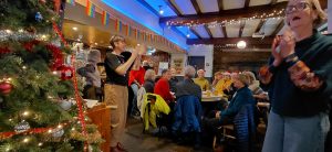Group of singers mostly sat at pub tables singing beside a Christmas tree