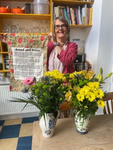 Clare is standing holding a tea towel with the words of Bread and Roses on , standing behind 2 vases of yellow and pink flowers