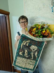 Rachael is standing holding a bouquet of beautiful yellow and orange flowers and a tea towel with words from John Ball of the Peasants Revolt